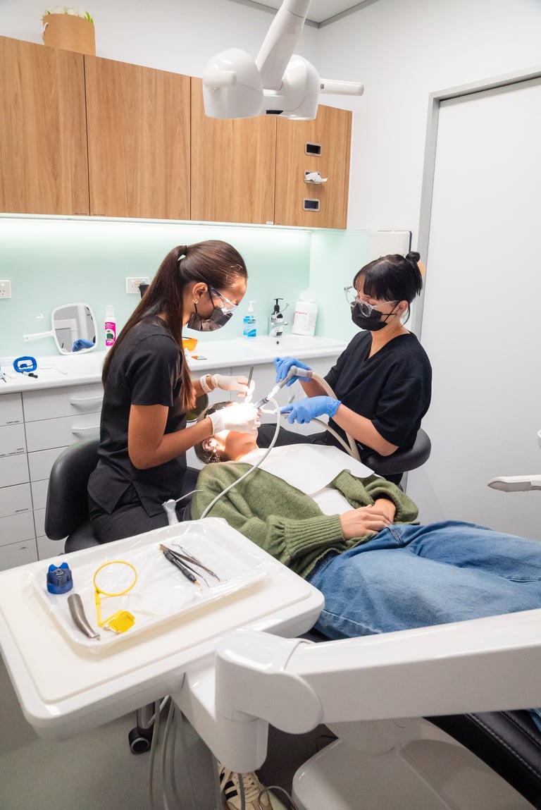 Two dental professionals in black masks and blue gloves working on a seated patient in a modern dental operatory with overhead lighting and wood cabinetry