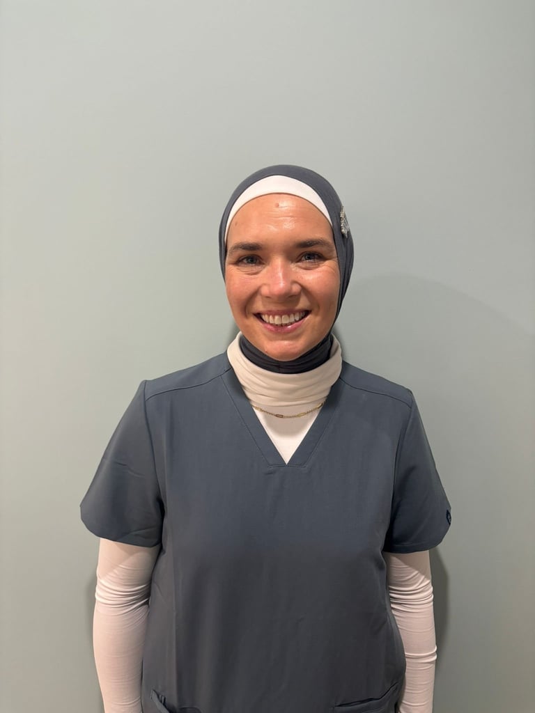 Portrait of smiling person wearing gray head covering, dark blue scrubs, and arm bandages against white background