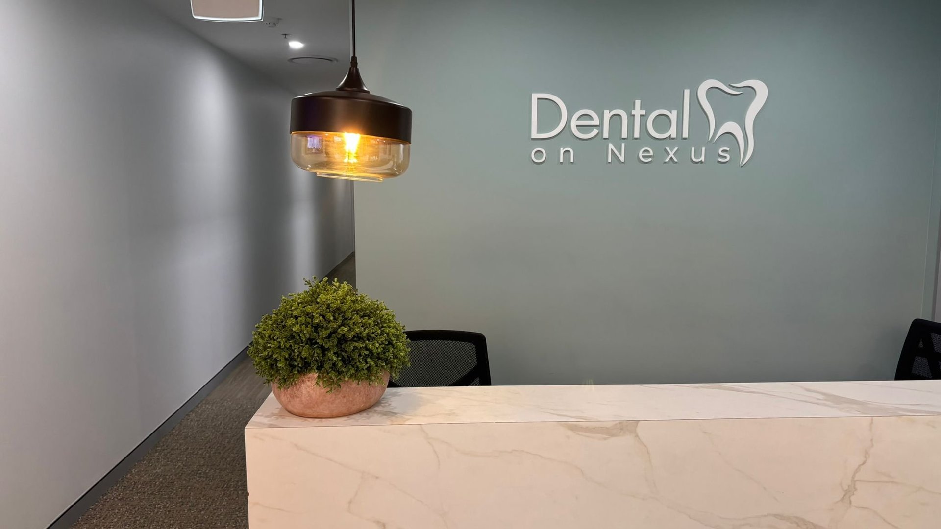 Modern dental office reception area with Dental on Nexus logo, marble desk, pendant lamp, and potted plant.