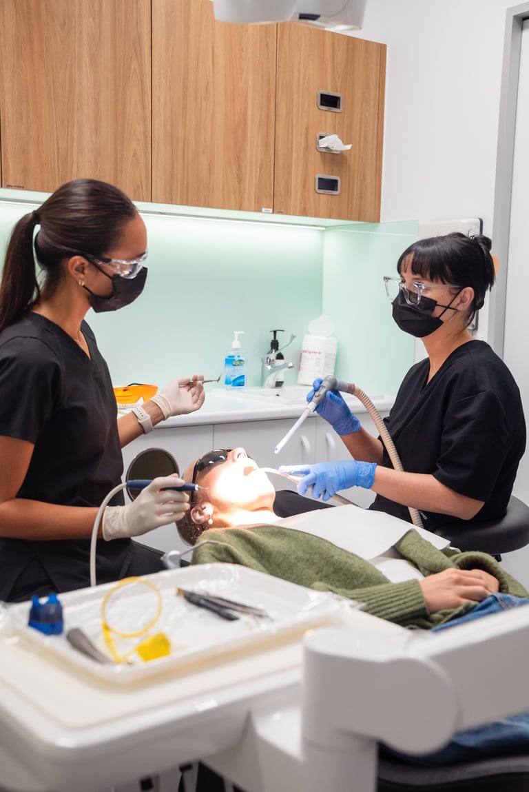 Two dental hygienists in masks and gloves treating a patient in a modern dental clinic with equipment and instruments visible